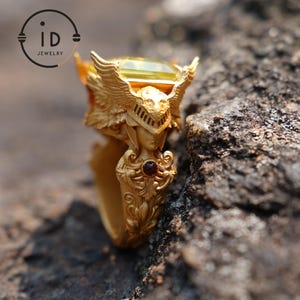 Puede incluir: Un anillo de oro con un diseño detallado de una figura alada que lleva un casco y sostiene una piedra preciosa. La piedra preciosa es de color amarillo claro y el anillo tiene una pequeña piedra preciosa roja en el lateral.