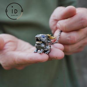 Puede incluir: Colgante de dragón plateado con ojos de piedras preciosas azules y cuernos dorados. El dragón está intrincadamente detallado y sujeto a una cadena de plata. El colgante se sostiene en las manos de una persona, mostrando la artesanía de la joyería.