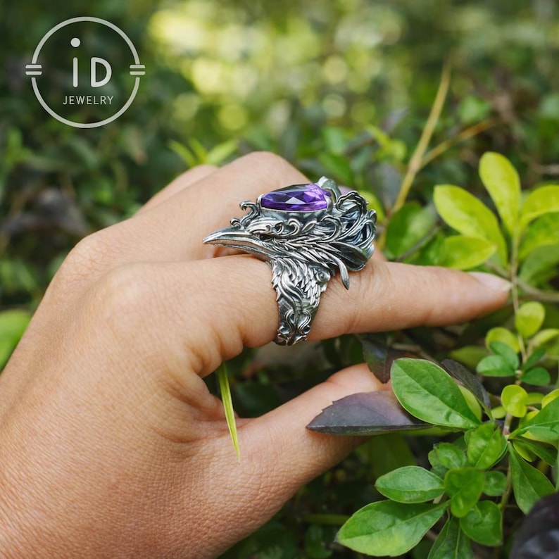 May include: A silver ring featuring an intricate, sculpted bird head design with detailed feathers and a prominent, pear-shaped purple gemstone set at the top. The metal has a dark, oxidized finish that highlights the textured relief work.