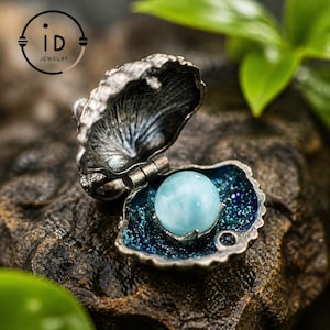 May include: A detailed shot of a silver-toned jewelry piece shaped like an open seashell. The interior is a deep blue with a light blue sphere. The jewelry has the text "iD JEWELRY" in a circular logo.