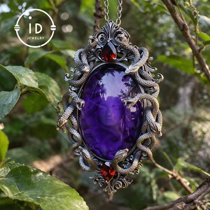 Könnte beinhalten: Eine silberfarbene Anhängerkette mit einem ovalen, violetten Stein, der mit einem Gesicht geschnitzt ist. Der Anhänger ist von kunstvollen Metallarbeiten mit gewundenen Schlangenmotiven und roten Edelsteinakzenten umgeben. Die Halskette hängt an einer zarten Kette.