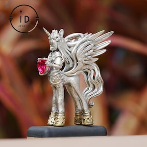 Peut inclure: Figurine de licorne argentée avec une crinière fluide, des ailes et une corne. La licorne tient une pierre précieuse rose en forme de cœur. La figurine a des accents dorés sur ses sabots. Le logo "ID Jewelry" est en haut à gauche.