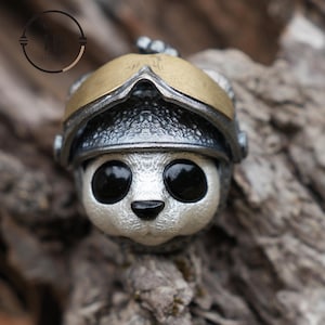 May include: A silver and gold metal figurine of a panda wearing a helmet with a visor. The panda has black eyes and a black nose.
