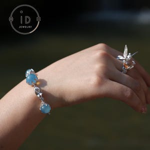 May include: A silver bracelet with three light blue gemstone beads and a matching ring with a brown gemstone and a dragonfly design. The jewellery is displayed on a person's hand.