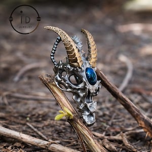 Op de afbeelding: Een zilverkleurige hanger in de vorm van een gehoornde schedel met een blauwe edelsteen oog. De hoorns zijn goudkleurig. De hanger wordt getoond op een takje tegen een vage natuurlijke achtergrond. Het ID Jewelry-logo staat linksboven.