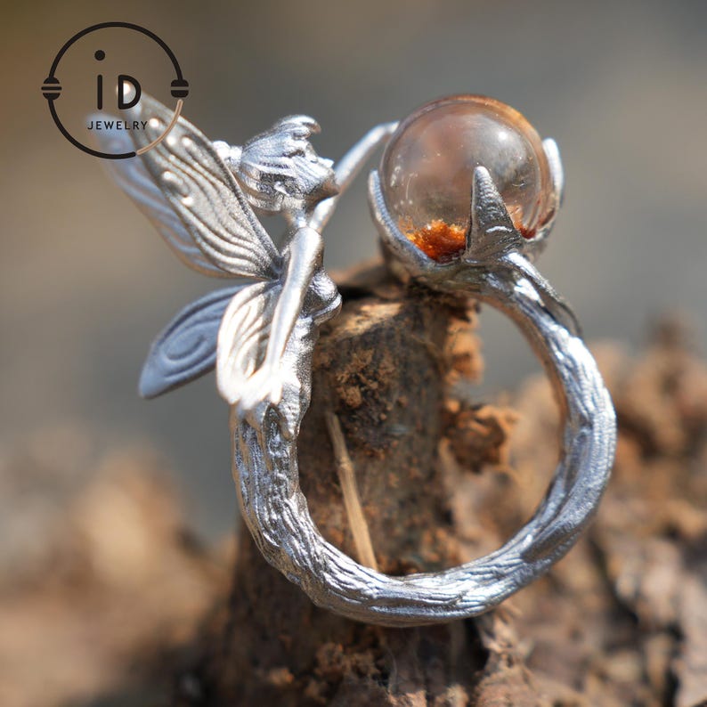 May include: A silver ring with a fairy motif. The ring's band has a textured, branch-like design, with a fairy figure and a clear orb holding amber-coloured inclusions. The ring is set against a natural wood background.