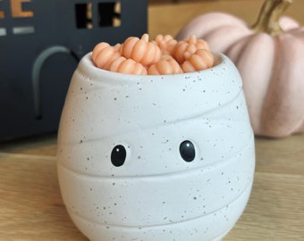 May include: A white ceramic mummy-shaped candle holder with black eyes and a speckled finish. The holder is filled with small, orange pumpkin-shaped wax melts.