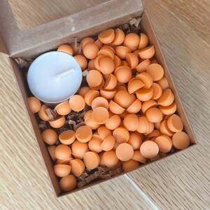 May include: A cardboard box with a clear window contains numerous orange wax melts and a white tea light candle. The wax melts are small, round, and a vibrant orange color. The box is filled with crinkle paper.