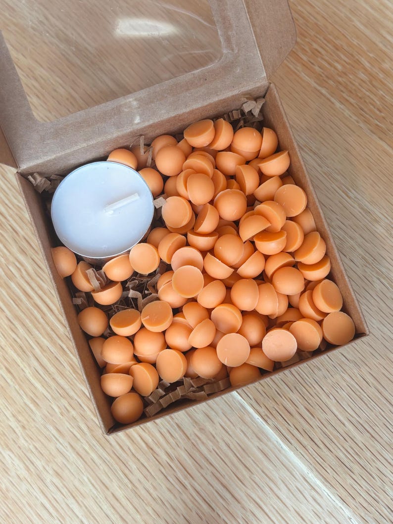 May include: A cardboard box with a clear window contains numerous orange wax melts and a white tea light candle. The wax melts are small, round, and a vibrant orange color. The box is filled with crinkle paper.