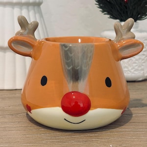 May include: A ceramic reindeer-shaped mug with an orange body, cream-colored base, and a red nose. The mug has black eyes, gray antlers, and a smiling mouth. A festive holiday decoration.