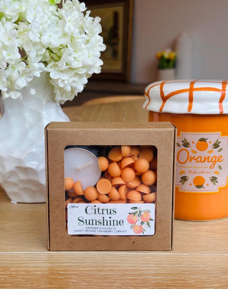 May include: A display of citrus-scented wax melts and a jar of orange marmalade. The wax melts are in a cardboard box with a clear window, labeled "Citrus Sunshine." The jar is orange with a white lid and orange grid pattern.