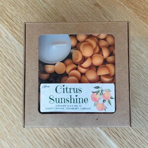 May include: A brown cardboard box with a clear window containing orange wax melts and a white tea light candle. The label reads "Citrus Sunshine" with the scent notes: sweet orange, cranberry, and currant.