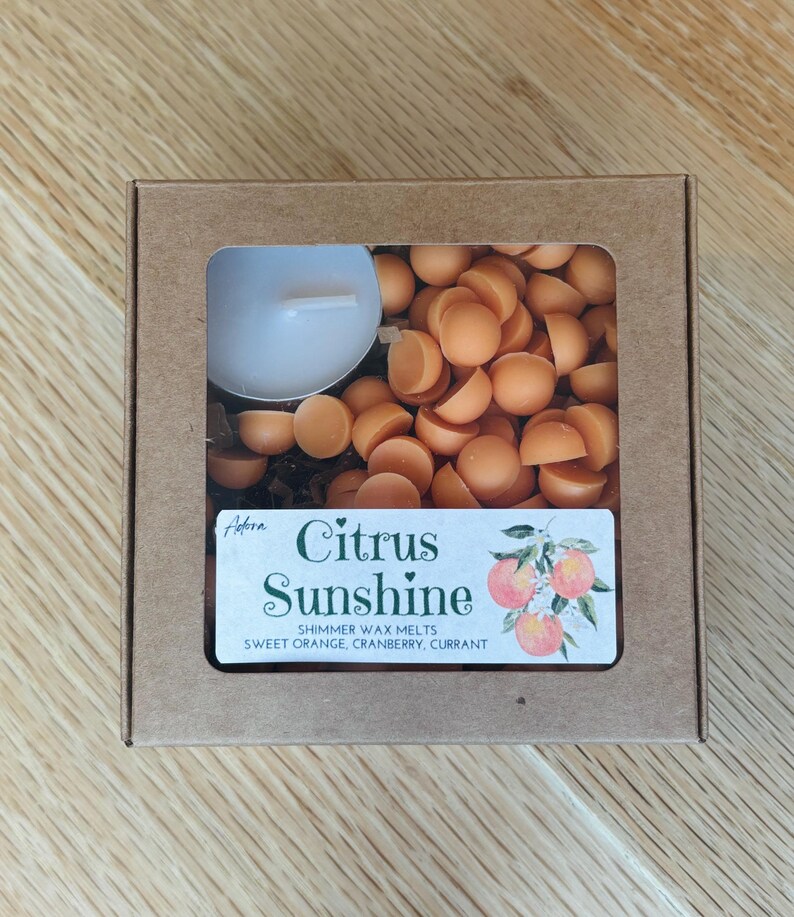 May include: A brown cardboard box with a clear window containing orange wax melts and a white tea light candle. The label reads "Citrus Sunshine" with the scent notes: sweet orange, cranberry, and currant.