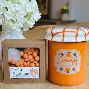 May include: An orange ceramic jar with a white and orange checkered lid, labeled "Orange Homemade Marmalade Made with love." Next to it is a box of "Citrus Sunshine" shimmer wax melts, featuring sweet orange, cranberry, and currant scents.