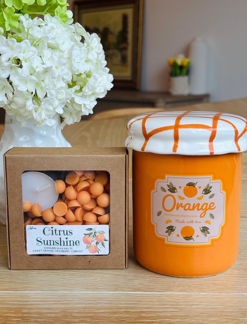 May include: An orange ceramic jar with a white and orange checkered lid, labeled "Orange Homemade Marmalade Made with love." Next to it is a box of "Citrus Sunshine" shimmer wax melts, featuring sweet orange, cranberry, and currant scents.