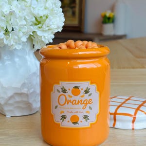 May include: An orange ceramic jar filled with orange-colored candies. The jar has a label that reads "Orange Homemade Marmalade Made with love." A white and orange checkered lid sits to the right.