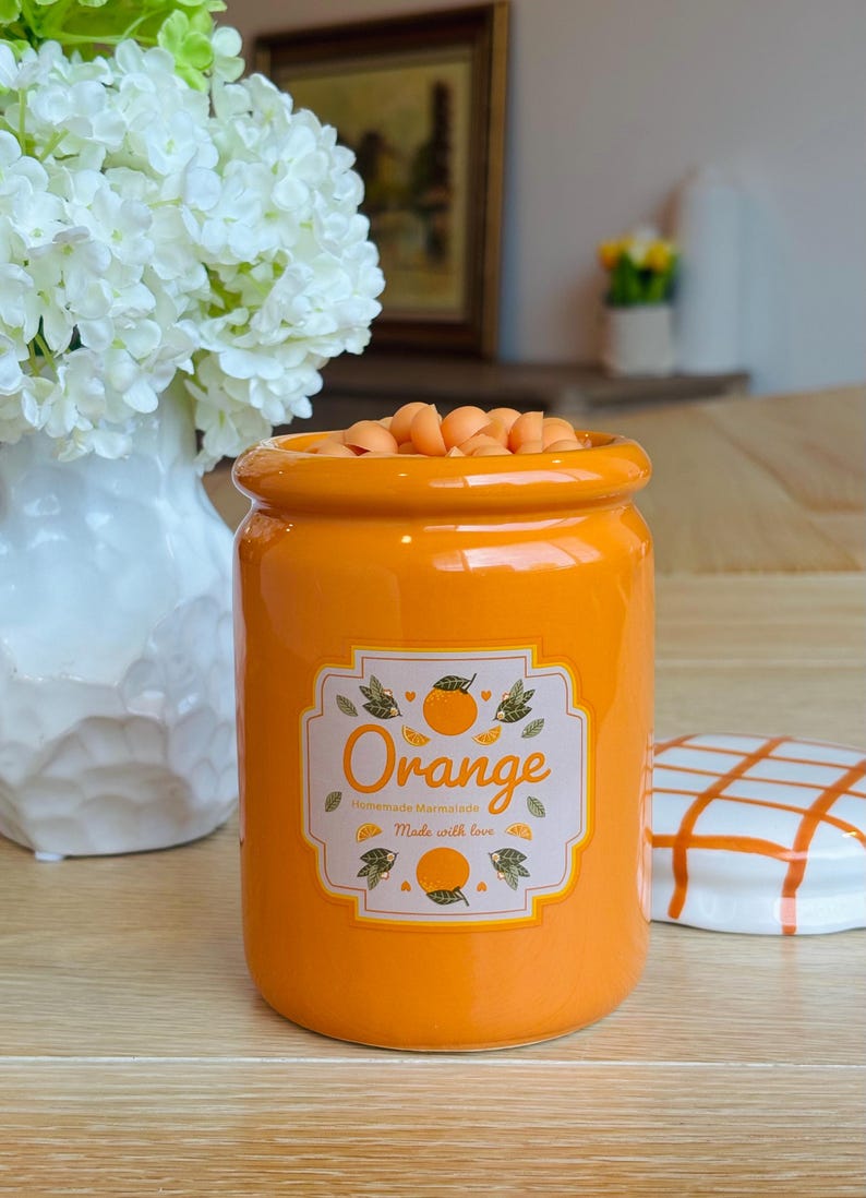 May include: An orange ceramic jar filled with orange-colored candies. The jar has a label that reads "Orange Homemade Marmalade Made with love." A white and orange checkered lid sits to the right.