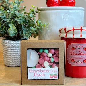 May include: A box of "Strawberry Patch" shimmer wax melts with a tea light candle. The box contains pink, white, and green wax melts. A jar of homemade strawberry jam and a white ceramic jar with strawberry-shaped lid are also visible.