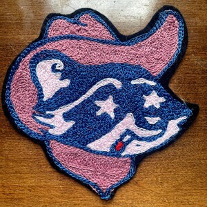 Op de afbeelding: Een geborduurde patch met een cartoonwasbeer die een cowboyhoed draagt. De wasbeer is roze en blauw met witte sterren op zijn voorhoofd en wangen.
