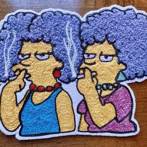 May include: Embroidered patch featuring two cartoon women with blue hair, yellow skin, and purple clothing. They are both smoking cigarettes and have a serious expression on their faces.