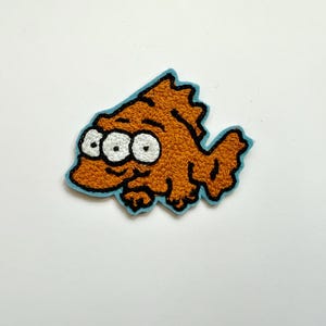 Könnte beinhalten: Ein gestickter Aufnäher mit einem Cartoon-Fisch mit drei Augen, in Braun mit einem blauen Umriss.