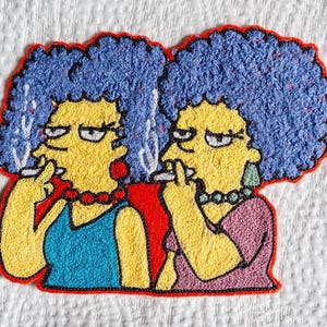 Puede incluir: Parche bordado con dos mujeres de piel amarilla y pelo azul, fumando cigarrillos. Llevan collares y pendientes, una con un top azul y la otra con un top rosa. El parche tiene un borde rojo.