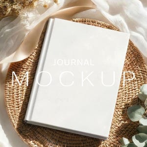 May include: A white journal mockup with the text "JOURNAL MOCKUP" on the cover, resting on a woven wicker tray. The scene includes dried flowers and a white fabric, creating a soft, natural aesthetic.