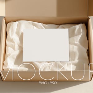 以下が含まれることがあります： 白いカードが中に入った長方形の開いた段ボール箱で、白い薄葉紙の上に置かれています。箱はベージュ色で、カードは白紙です。「MOCKUP」の文字が箱の下部に細い白いフォントで書かれており、その下に「PNG+PSD」と書かれています。
