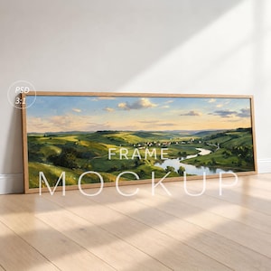 Op de afbeelding: Een panoramisch landschapsschilderij in een lichthouten lijst met glooiende groene heuvels, een kronkelende rivier en een klein dorp onder een zachte zonsonderganghemel. Over het kunstwerk staat de witte tekst PSD 3:1 FRAME MOCKUP.