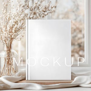 May include: A blank white book stands upright on a wooden surface, with the word "MOCKUP" faintly visible. A glass vase with dried flowers and a white fabric drape add to the minimalist aesthetic, set against a window.