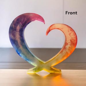 May include: A colorful heart-shaped sculpture with a gradient of colors, including pink, blue, yellow, and orange. The word "Front" is visible on the right side of the sculpture. The sculpture is made of a translucent material and sits on a wooden surface.