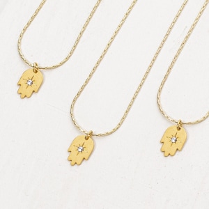 May include: Three gold necklaces with a hamsa hand charm pendant featuring a small clear rhinestone. The hamsa hand is a symbol of protection and good luck.