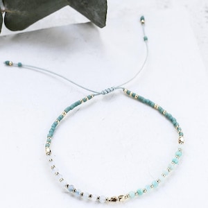 May include: A delicate adjustable bracelet featuring a mix of small teal, light blue, and white beads interspersed with tiny metallic gold-toned accents. The bracelet is strung on a thin, light blue cord with a sliding knot closure mechanism.