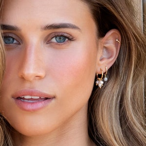 May include: A woman wearing gold hoop earrings with pearl accents. The earrings are layered with a smaller hoop and a single pearl dangle. The woman is also wearing a pearl necklace.