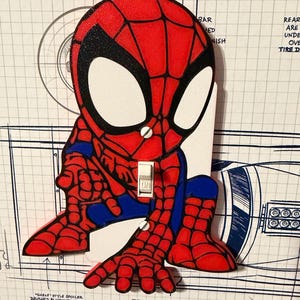 Könnte beinhalten: Eine rote, blaue und schwarze Spider-Man-Lichtschalterabdeckung. Die Abdeckung zeigt eine Cartoon-Darstellung von Spider-Man mit einem weißen Lichtschalter in der Mitte. Der Hintergrund hat blaue technische Zeichnungen und Text.