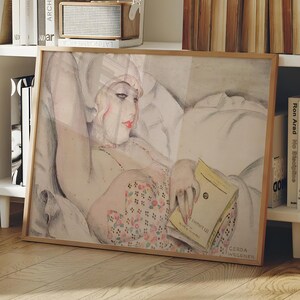 以下が含まれることがあります： 本を読んで横たわっている女性を描いた額入りのアートワーク。アートワークは、明るい茶色の木製フレームです。女性は花柄の衣服と白い帽子を着用しています。アートワークは白い棚に飾られています。