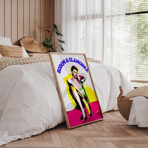May include: A framed art print featuring a stylized illustration of a person in a pose, with the text "SODOM & GLAMORRA" in blue. The artwork uses vibrant colors, including pink, yellow, and green, set against a white and gray background.
