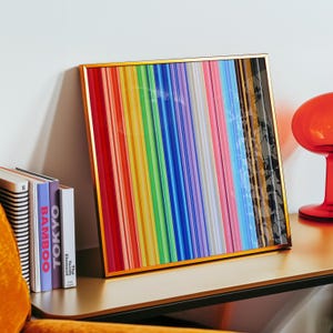Puede incluir: Una obra de arte enmarcada con un borde dorado presenta un vibrante arcoíris de rayas verticales. La obra de arte se apoya contra una pared blanca sobre una mesa dorada, junto a una pila de libros y un objeto de vidrio rojo.