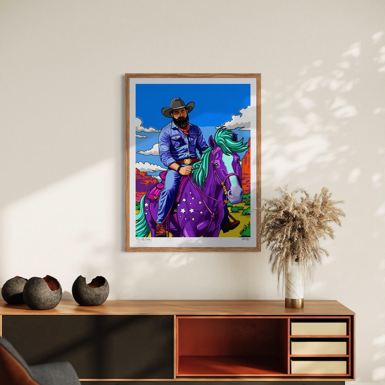 May include: A framed art print featuring a cowboy riding a purple horse with a starry pattern. The cowboy wears a blue shirt and hat, set against a desert landscape with a blue sky and clouds. The artwork has vibrant colors and a pop art style.