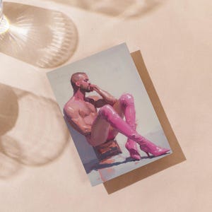 May include: Art print depicting a nude figure in pink boots, seated and leaning. The artwork is on a white background, placed on a brown card. A glass of water is in the upper left corner, casting a shadow across the surface.