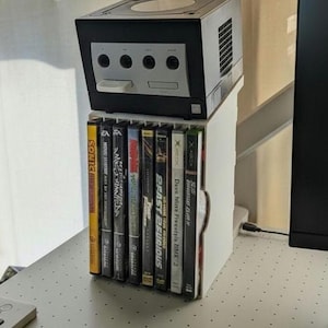 May include: A white and black video game console with a stack of game cases. The console has a rectangular shape with a circular top and several buttons. The game cases are in a vertical row.