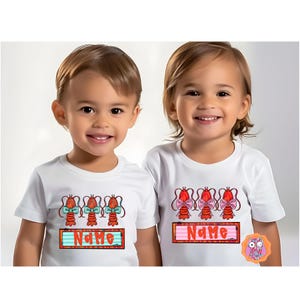 May include: Two white t-shirts featuring a design of three red lobsters wearing bows. The lobsters on the left shirt have blue bows and the lobsters on the right shirt have pink bows. The shirts have a red, white and blue striped banner with the word "NAME" printed on it.