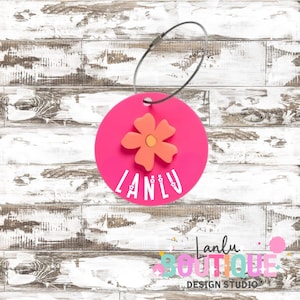 May include: A vibrant pink luggage tag featuring an orange flower and the white text "LANLU". The tag is circular and connected to a silver metal loop. Set against a backdrop of white-painted wooden planks.