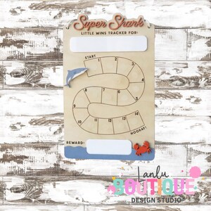 May include: Wooden "Super Shark" little wins tracker board with a shark and crab accent. The board has a path with numbered spaces, a "Start" and "Reward" section, and the text "Little Wins Tracker For:" and "Hooray!"