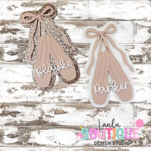 May include: Two pairs of ballet slippers in a soft pink hue, each with a delicate bow and ribbons. One pair is outlined with glitter and has the name "Blayke" in white script. The other pair has the name "Parker".
