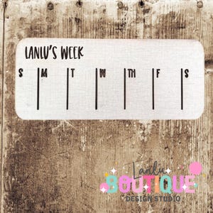 May include: A white rectangular weekly planner with the text "LANLU'S WEEK" at the top. The days of the week are abbreviated below the text, with vertical lines for each day. The planner is set against a wooden background.