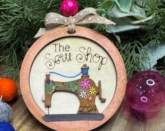 May include: A circular wooden Christmas bauble with a brown border. The bauble features a stylised illustration of a sewing machine with colourful flowers and the text "The Sew Shop 2024".