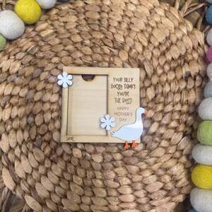 May include: A square wooden photo frame featuring a white goose cutout with an orange beak and feet, two white flower embellishments, and the text "Your silly goose thinks you're the best! Happy Mother's Day" printed in black ink on the right side.