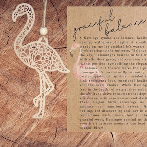 May include: A laser-cut wooden flamingo ornament with an intricate filigree pattern, suspended by a jute string with a single wooden bead. Beside it, a rectangular card features the text "graceful balance" and a printed illustration of a pink flamingo.