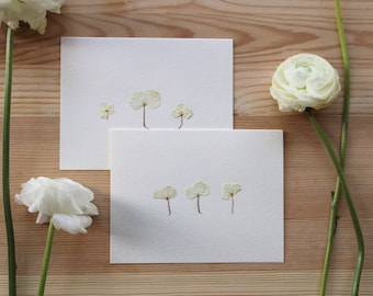 Peut inclure: Deux cartes blanches avec des fleurs vertes pressées disposées en motif. Les fleurs sont disposées en rangée sur chaque carte. Les cartes sont sur une surface en bois brun clair.